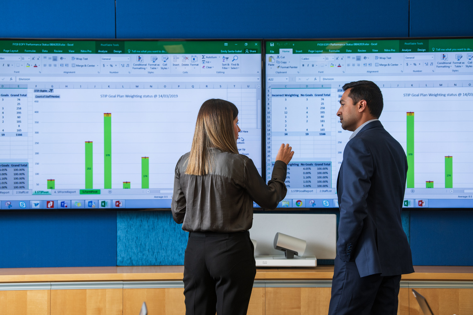 A man and a woman in an office talking in front of large presentation screens